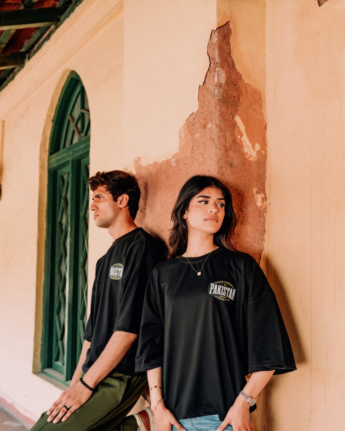 Two models in Zamana Alumni Tees against an ochre wall beside a green shuttered window, Lahore