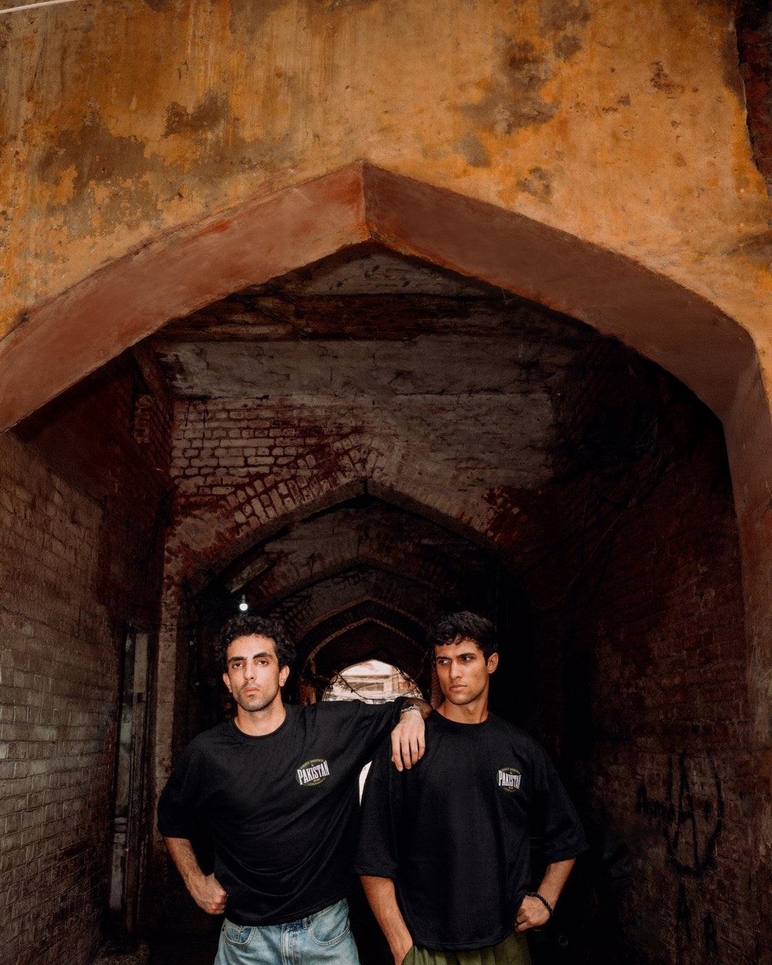 Two models in Zamana Alumni Tees framed by a brick archway in Lahore