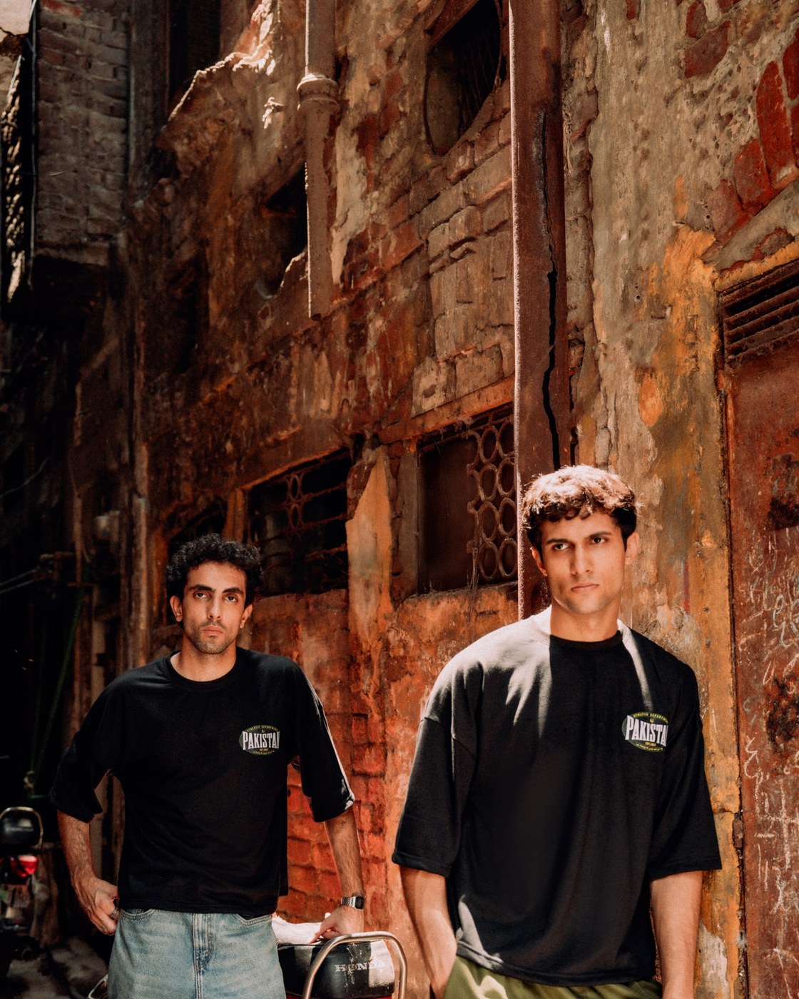 Two models in Zamana Alumni Tees on a Lahore street, brick and shuttered facades behind them