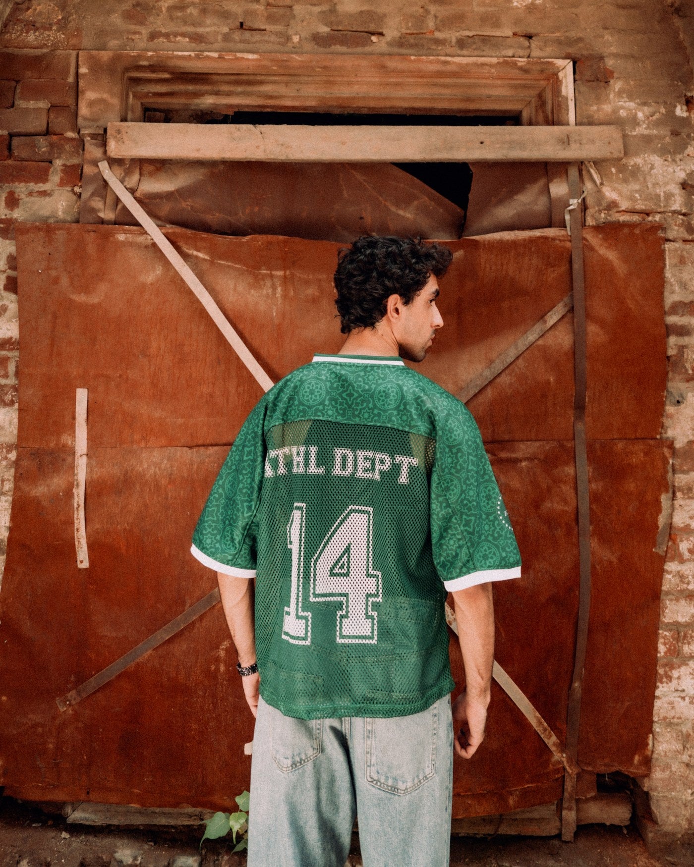 Dark green Athletic Department jersey worn from behind, ATHL DEPT 14 across the back, before a red door in a brick doorway in Lahore