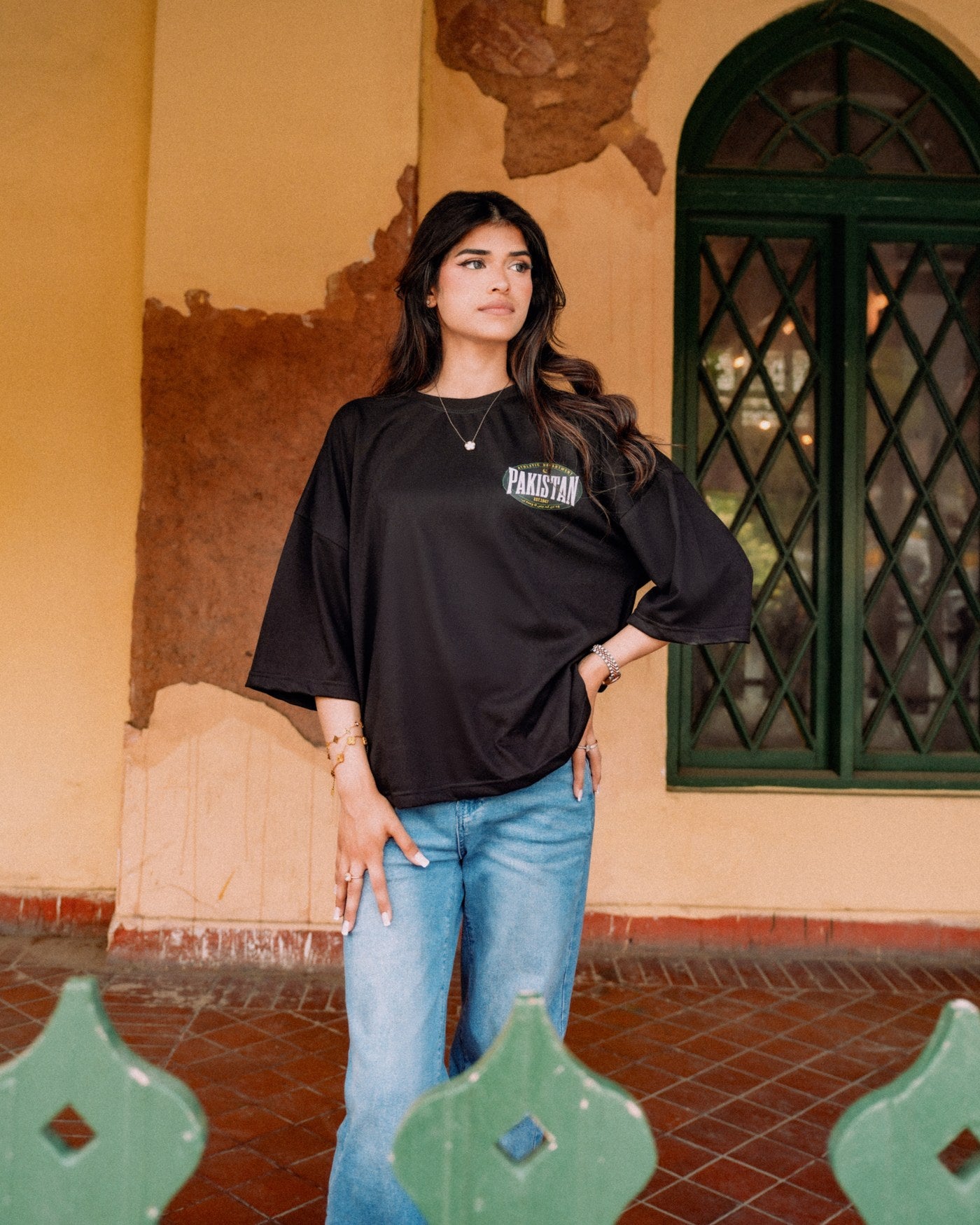 Model in a black Athletic Department PAKISTAN tee against an ochre veranda wall and green lattice window, shot on location in Lahore